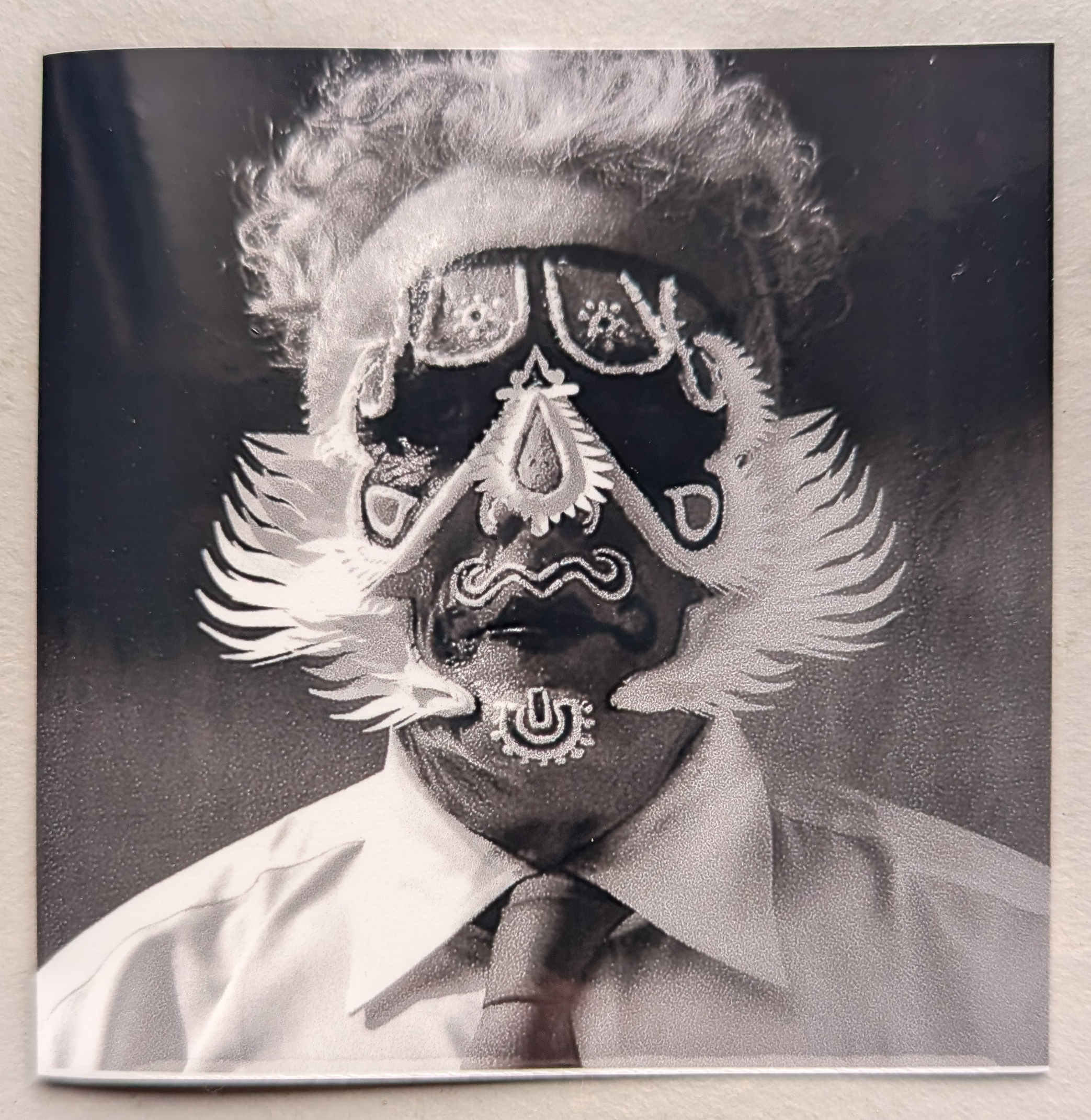 A black and white portrait of a person wearing a decorative mask with intricate designs, featuring feathers and unique patterns, against a blurred background.