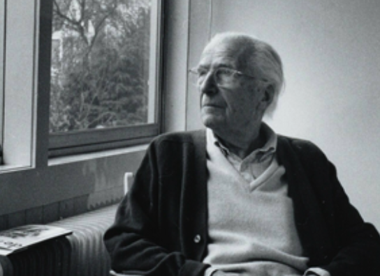 An elderly man with glasses sits thoughtfully by a window, looking out at the view. He is wearing a cardigan over a light sweater and appears to be in a cozy indoor setting.